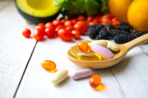 Pills & Capsules in wooden spoon with fruits & veggies in background.